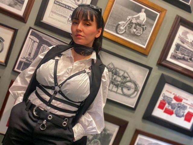 SaraleePoison with black strappy harness, white button-up shirt, black pants, and black choker, standing in front of motorcycle-themed wall art. She has medium tits and straight black hair.