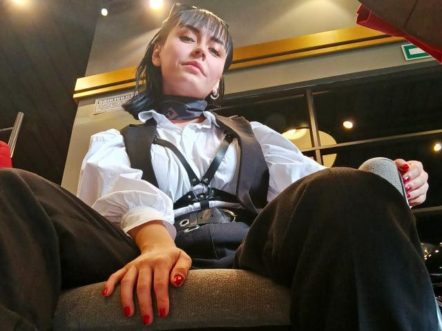 SaraleePoison with black hair, white shirt, black suspenders, and black pants, sitting with legs apart, holding a red-cupped drink, red nails, and a confident, slightly smug expression.