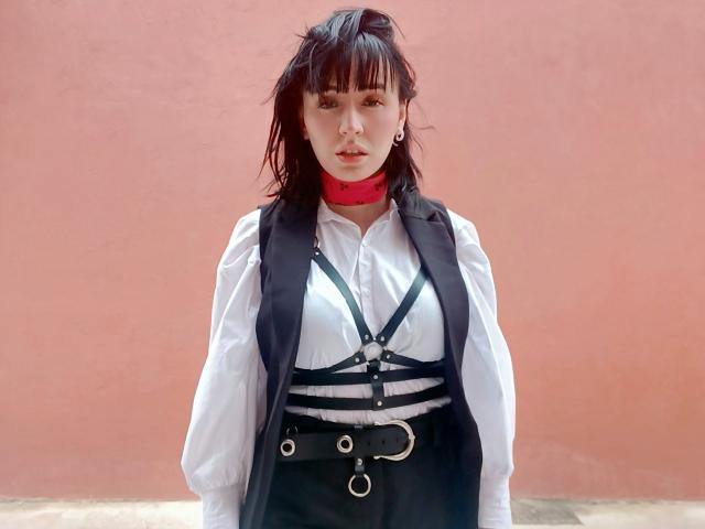SaraleePoison with dark wet hair, red choker, white shirt, black harness, and straps, black vest, belt with rings, standing against a pink wall, looking forward.