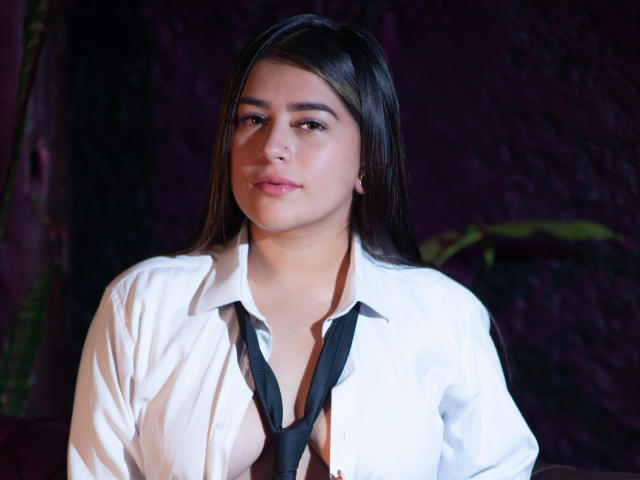 Nicole Acosta, met lang bruin haar, draagt een licht open wit shirt en een zwarte stropdas, waardoor haar kleine, stevige borsten zichtbaar zijn. Ze kijkt recht in de camera met een neutrale uitdrukking..