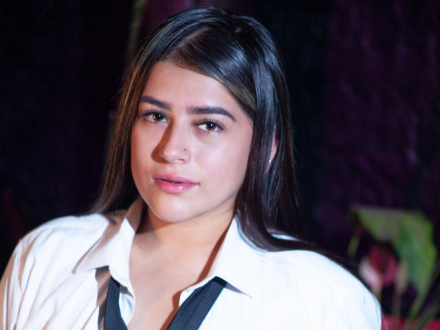 Nicole Acosta, met lang donker haar, die een witte blouse draagt, zwarte braces en een kleine neusring, heeft een licht blozende, schattige gezicht en subtiele, roze lippen..