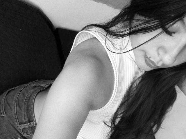 Fotografia in bianco e nero di ClaraPattel con lunghi capelli scuri, labbra piene, che indossa una canotta bianca a coste e jeans, mostrando il suo braccio e fianco, con un viso leggermente sfocato..