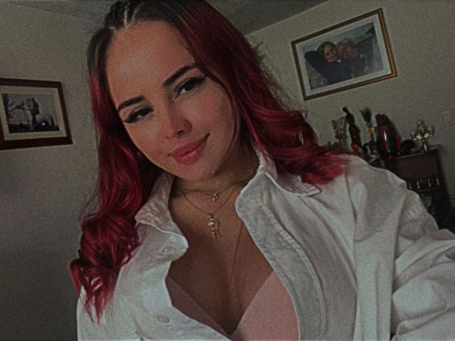 Nia Spencer, con cabello rojo, lleva una camisa blanca abierta que revela su sujetador rosa y su escote. Está sonriendo, lleva un collar y tiene un aspecto lindo y atractivo..
