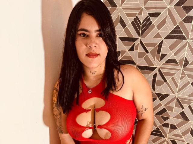 Mandy, kurvig, mit schwarzen Haaren, einem roten Netzoberteil, das ihre großen Brüste freilegt, Tattoos an den Armen und einem kleinen Ring an der Brustwarze, steht vor einer Wand mit geometrischen Mustern..