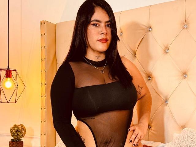 Mandy, con lunghi capelli neri, indossa una maglia nera trasparente a maniche lunghe, che mostra il seno e un tatuaggio sul braccio destro, ed è in piedi di fronte a una testiera beige trapuntata..