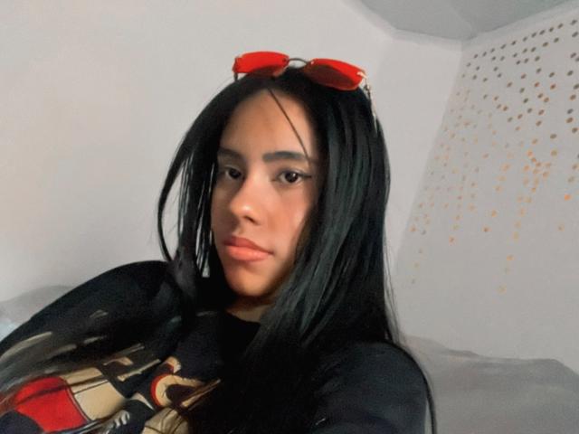 SiennaTaylor has long black hair, red sunglasses on her head, and a serious look on her face, wearing a black shirt with red and gold patterns.