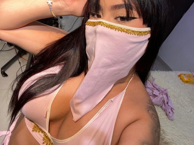 MiaHadid with dark hair, pink face mask, and pink top showing big tits, gold trim, arm raised, visible tattoo, and bracelet. Her eyes are lined with black eyeliner.