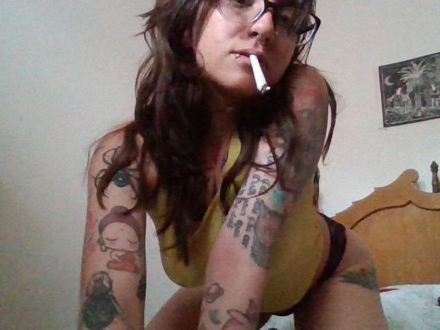 AmySpider, met lang bruin haar, een bril en een sigaret in haar mond, leunt voorover in een geel tanktopje, waarbij meerdere tatoeages op haar armen en benen zichtbaar zijn..