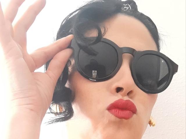 Claudias with black round sunglasses, red lipstick, and black curly hair, adjusting her glasses with her right hand, showing off her gold earrings.