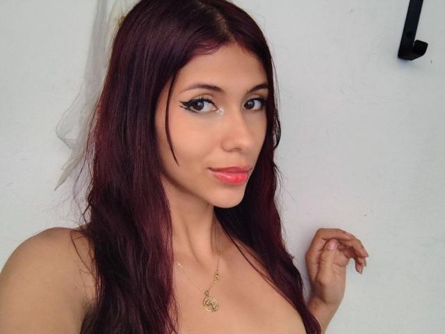 DanikaSerov with dark red hair, blue eyes, and pink lipstick, wearing a gold necklace, showing her tits, and a slight smile, against a white wall.