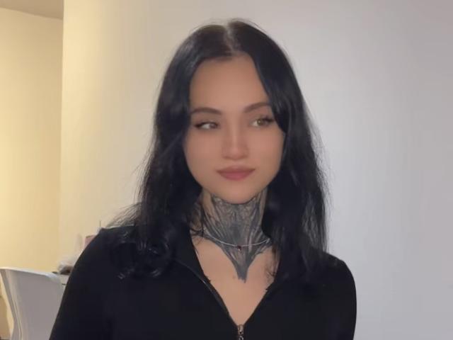 BlackInkQueen with black wavy hair, tattoos on her neck, wearing a black zip-up, staring off to the side with a slight smile, pale skin, and subtle makeup.