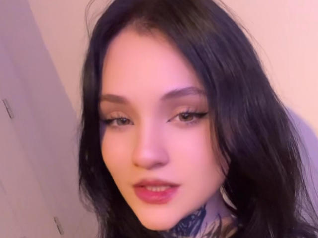 BlackInkQueen has long black hair, light skin, and eyeliner. She's looking at the camera with a slight smile, showing her pink lips and a tattoo on her neck.