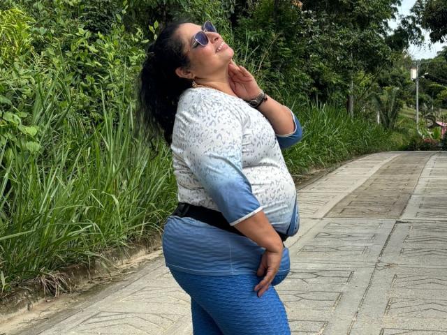 Keyla69Hot, chubby with a big ass, wearing a white and blue patterned top, blue quilted pants, and blue sunglasses, stands on a sidewalk with greenery behind her.