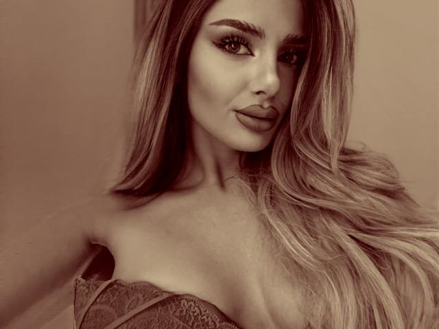 RheaI has long, wavy blonde hair, full lips, and is wearing a lace strapless top that shows her large tits. Sepia-toned, sexy, confident.