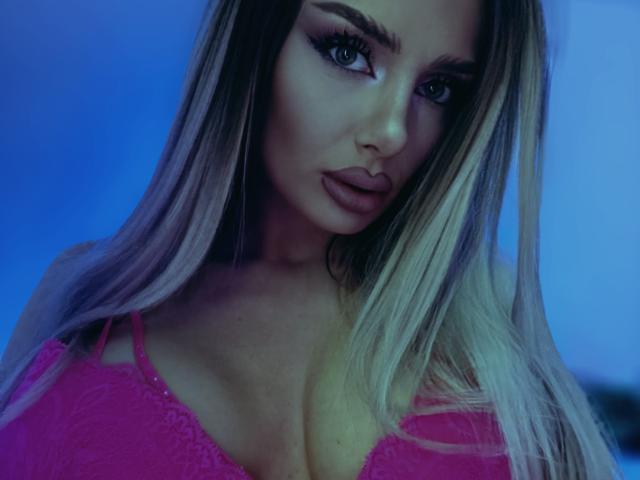 RheaI has blonde hair, blue eyes, and wears a pink lace bra, showing her big tits and pouty lips, with a blue-tinted background.