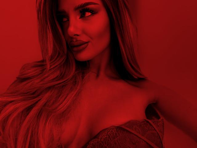 RheaI has red-lit skin, long blonde hair, and a sultry smile, wearing a lace bralette that shows her tits. The image is red and sexy.