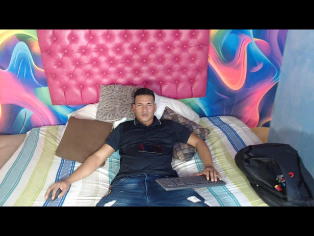 JeicodLatino with short black hair, wearing a black shirt and blue jeans, lying on a bed with a pink tufted headboard, holding a keyboard, laptop on his lap.