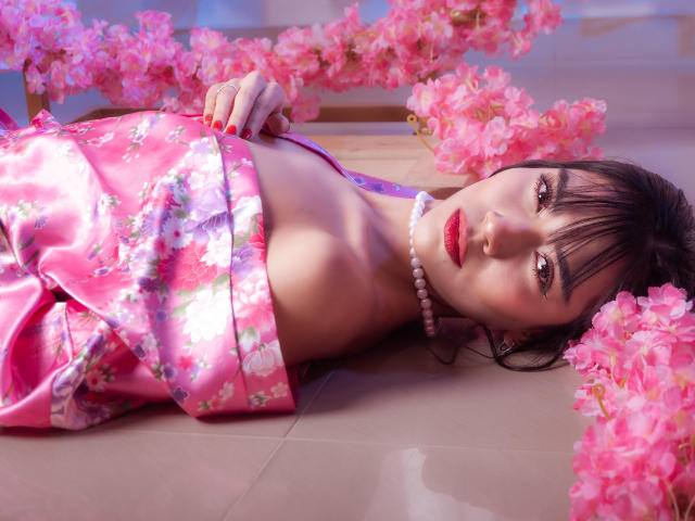 Ein Engel mit rosafarbenem Kimono mit Blumenmuster, liegt auf einem Fliesenboden, umgeben von rosa Blumen. Sie hat roten Lippenstift, eine Perlenkette und schwarzes Haar mit Pony und blickt in die Kamera..