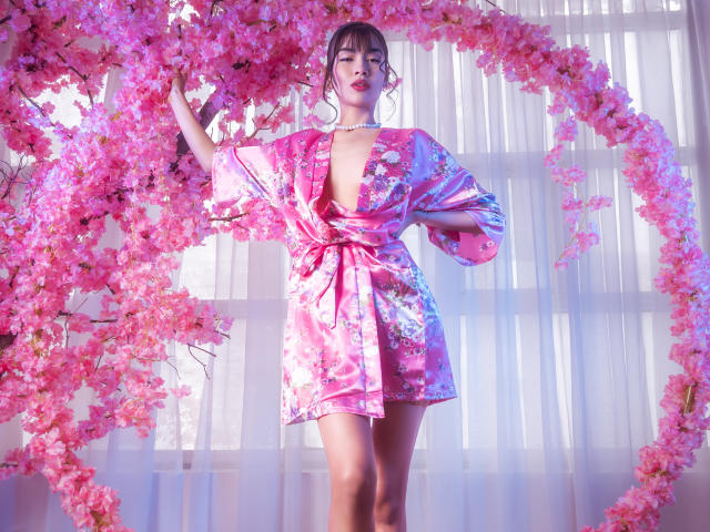 TinnyEngel, vestindo um quimono floral rosa que expõe seus seios, está em pé sob um arco de cerejeiras com flores rosa, usando um colar de pérolas, com a mão levantada..