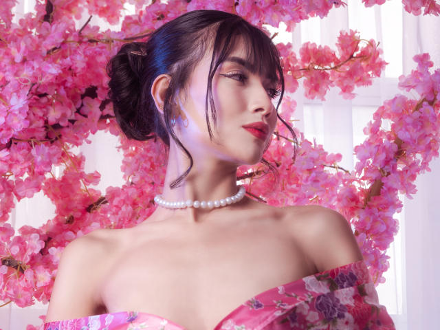 TinnyEngel con sfondo di fiori di ciliegio rosa, indossa una maglietta rosa a fiori con una spalla scoperta, una collana di perle e rossetto rosso, guarda di lato, con capelli scuri raccolti in uno chignon. Il seno è visibile..