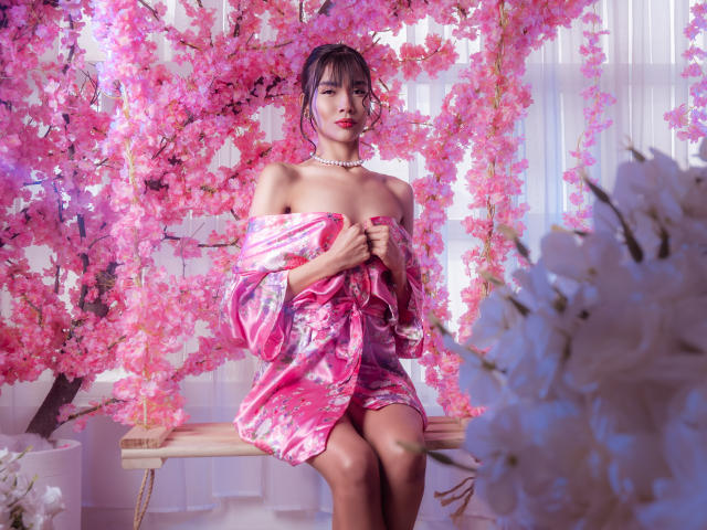 TinnyEngel mit einem pinken Kimono mit Blumenmuster, schulterfrei, der kleine Brüste zeigt, sitzt auf einer hölzernen Schaukel, umgeben von rosa Kirschblüten, trägt eine Perlenkette, rote Lippenfarbe und hat schwarzes Haar..
