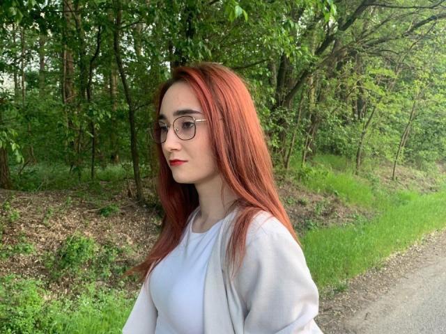ChaudesChatteX has red hair, glasses, and red lipstick, wearing a white shirt, standing by a grassy path with a forest in the background. Her tits are small.