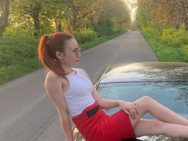 ChaudesChatteX, with red ponytail and glasses, sits on a car's roof, wearing a white tank top and red skirt, showing her ass, in a sunlit, wooded road.