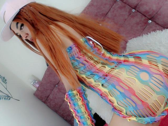 Kendalla, con cabello largo y naranja, usando un sombrero blanco, con un traje de baño colorido y de una sola pieza, que deja ver su trasero, de pie frente a una cama rosa con volantes..