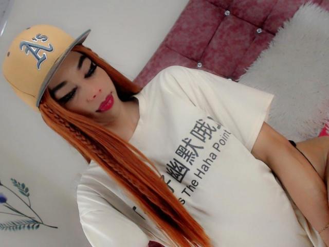 Kendalla mit langen, orangefarbenen Zöpfen, die eine gelbe Mütze, ein weißes T-Shirt und roten Lippenstift trägt, zeigt ihr süßes Gesicht und ihren kleinen Hintern. Sie hat dunkle Augenbrauen und volle, rosafarbene Lippen..