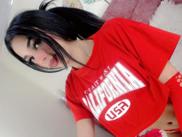 Kendalla with black hair, wearing a red California USA T-shirt, showing her midriff and a small nose piercing, sitting on a pink fluffy chair. Boobs visible, cute face.
