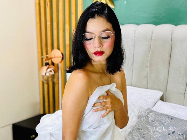 AngelicMilers met zwart haar, rode lippenstift en een bril, die een witte handdoek over haar blote borsten houdt, en naar beneden kijkt met een lichte blos. Ze ligt op een bed met grijze kussens..