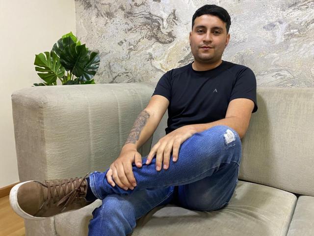 AlhanOliver, con cabello corto y negro, camiseta negra, jeans azules rotos, botas marrones y un tatuaje en el brazo derecho, sentado en un sofá gris, mirando a la cámara, con las manos sobre las rodillas..