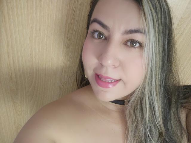 AnmeLeon with pink lips, brown eyes, nose piercing, black choker, and blonde hair, smiling at the camera, showing her bare shoulders.
