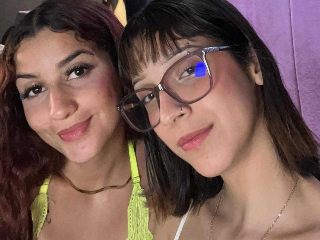 Ktita with curly red hair and small tits, wearing a neon yellow top, stands next to Lia with black hair, glasses, and a nose ring, smiling at the camera.