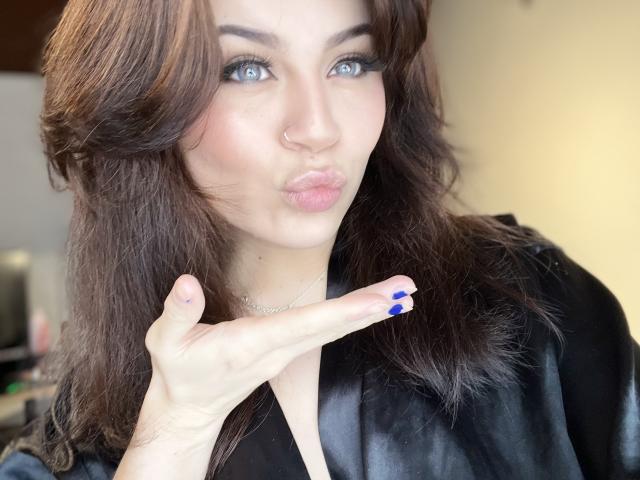 ShantallParis, con le unghie blu, le labbra carnose e un piercing al naso, indossa un vestito di seta nera, ha i capelli castani ondulati e gli occhi azzurri, e fa una smorfia da bacio..