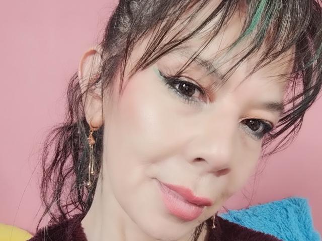 SamaraBellet's face with pink lips, black eyeliner, and blue-green hair streak, wearing earrings, smiling softly, against a pink background, showing a cute and attractive appearance.