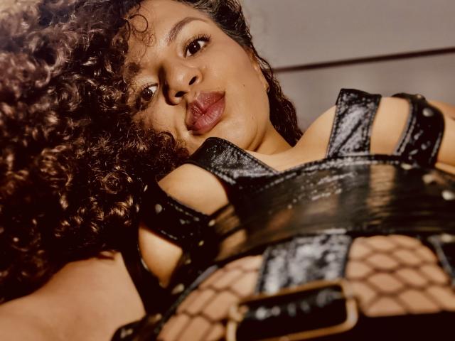TiaraKitty with curly brown hair, black leather strappy top, and fishnet bottom, pouting her full, glossy red lips, wearing a shiny black belt, in a low-angle shot.