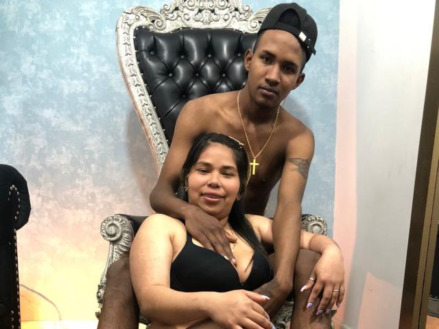 LucasAndLily, shirtless with a gold cross necklace, sits on an ornate black chair, arms around a chubby woman with black tits in a black bra, wearing a backwards cap.