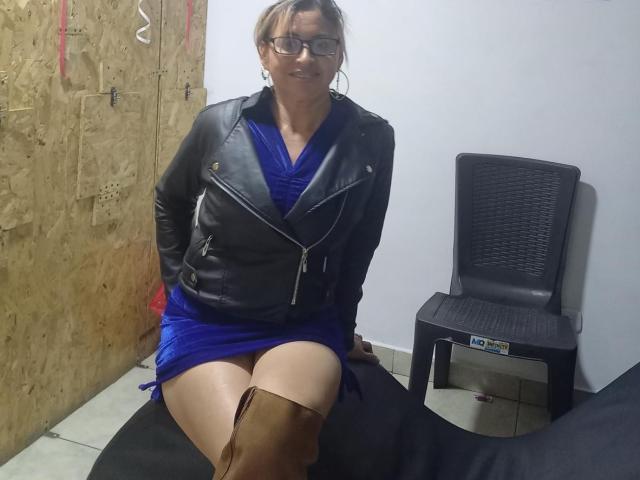 SherahaJade sentada con el trasero expuesto, vistiendo un vestido azul, una chaqueta de cuero negro y botas marrones hasta las rodillas, sonriendo, con gafas, y con las manos sobre los muslos..