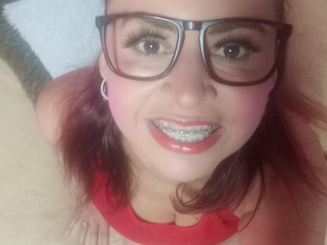SherahaJade, con gafas, lápiz labial rojo y dientes con brillo, sonriendo a la cámara. Lleva una blusa roja y tiene colorete rosa en las mejillas, con cabello castaño..