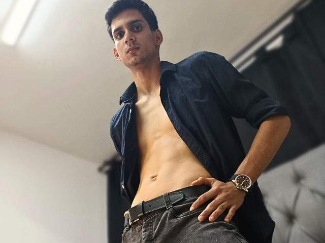 SebaLexis, con un torso asciutto e muscoloso, senza camicia, indossa una camicia blu scuro, pantaloni grigi e un orologio, ed è in piedi con le mani sui fianchi, guardando in basso..