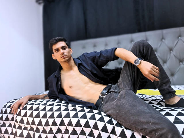 SebaLexis, ohne Hemd, mit einem schlanken, trainierten Oberkörper, einer schwarzen Uhr und dunklen Jeans, liegt auf einem schwarz-weiß gemusterten Bett und blickt mit ernstem Gesichtsausdruck in die Kamera..
