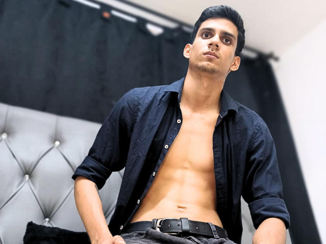 SebaLexis, con la camicia slacciata che lascia scoperto un petto muscoloso e tonico, una cintura nera e pantaloni neri, guarda in basso con un'espressione seria, ha i capelli scuri e la pelle di media tonalità, e si trova in una camera da letto..