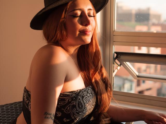 AnniaLeRoux with long red hair, wearing a black hat and strapless black and white patterned top, sunlight on her face, tattoo on right arm, eyes closed, smiling.