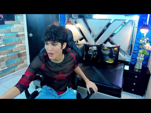 MarcoMiranda, short black hair, red and black sheer shirt, blue jeans, sitting in black leather chair, looking annoyed, mirror with lights, black bag, flower vase on right.