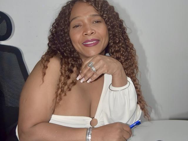 AinaEbonyQueen with curly brown hair, smiling, wearing a white dress with a keyhole, showing her tits, holding a blue pen, ring on finger, and silver bracelet.