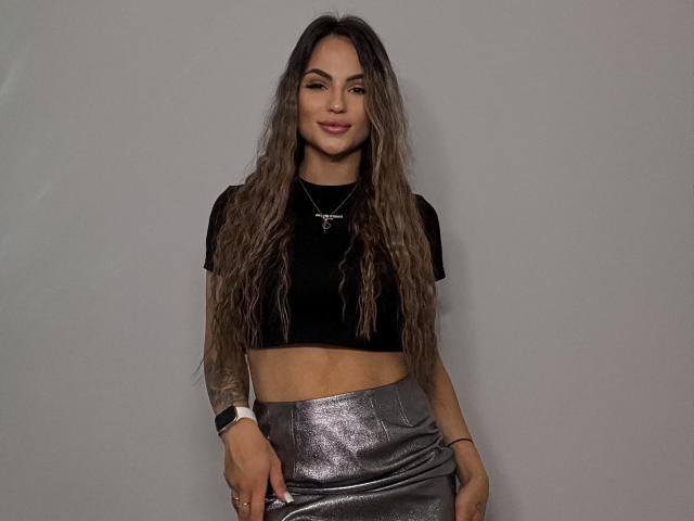 KarolinaSykes with long wavy brown hair, black crop top, silver shiny skirt, and a necklace, standing, smiling, wearing a smartwatch on her left wrist.