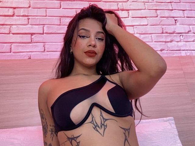 ValentinaMathews with long dark hair, wearing a black one-shoulder bra, showing tattoos on her chest and arm, hand in hair, red lipstick, and a confident expression.