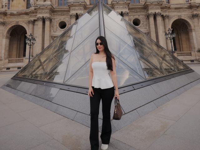 ArianaGildmore stands in front of a glass pyramid, wearing a white strapless top, black flared pants, and white heels, holding a brown handbag, with long black hair.