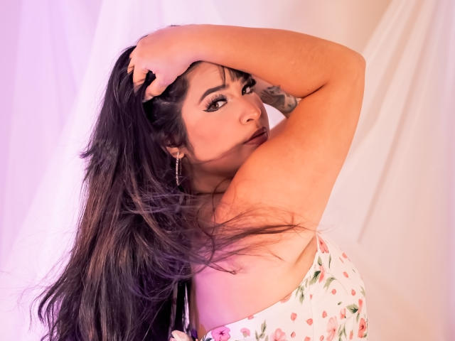 AntonelaPark with long dark hair, hand in hair, looking over shoulder, floral dress, pinkish-purple lighting, tattoos visible on arm, earrings, soft expression, slightly parted lips.
